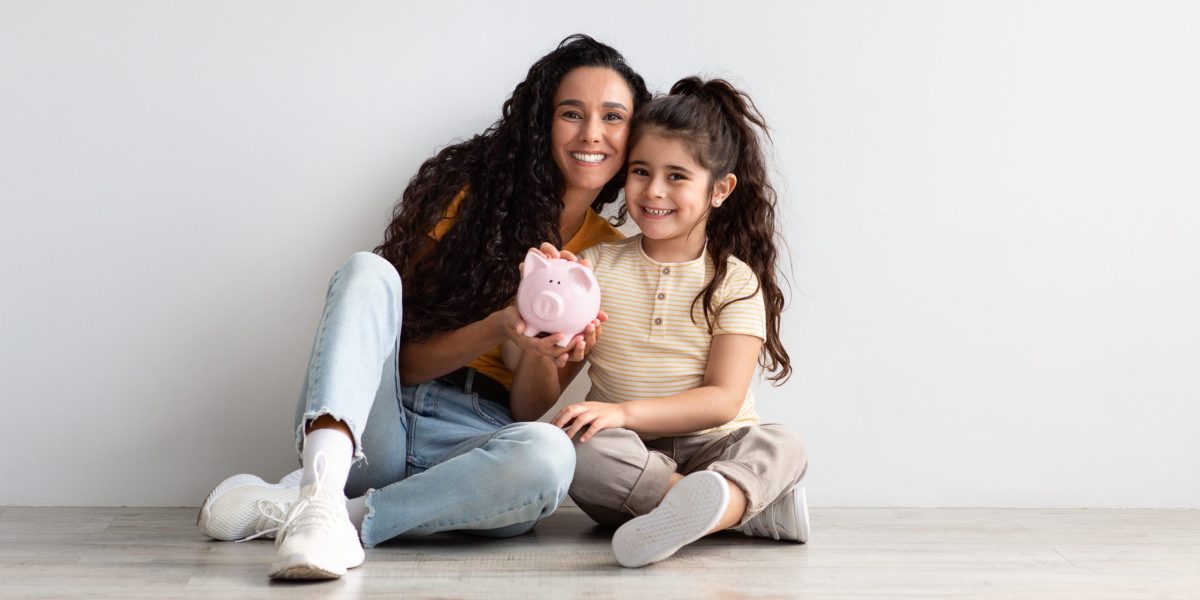 Financial Literacy. Portrait Of Young Mom And Her Little Daughter With Piggybank In Hands Sitting On Floor Near White Wall At Home, Smiling Mother Teaching Her Child Saving Money, Copy Space